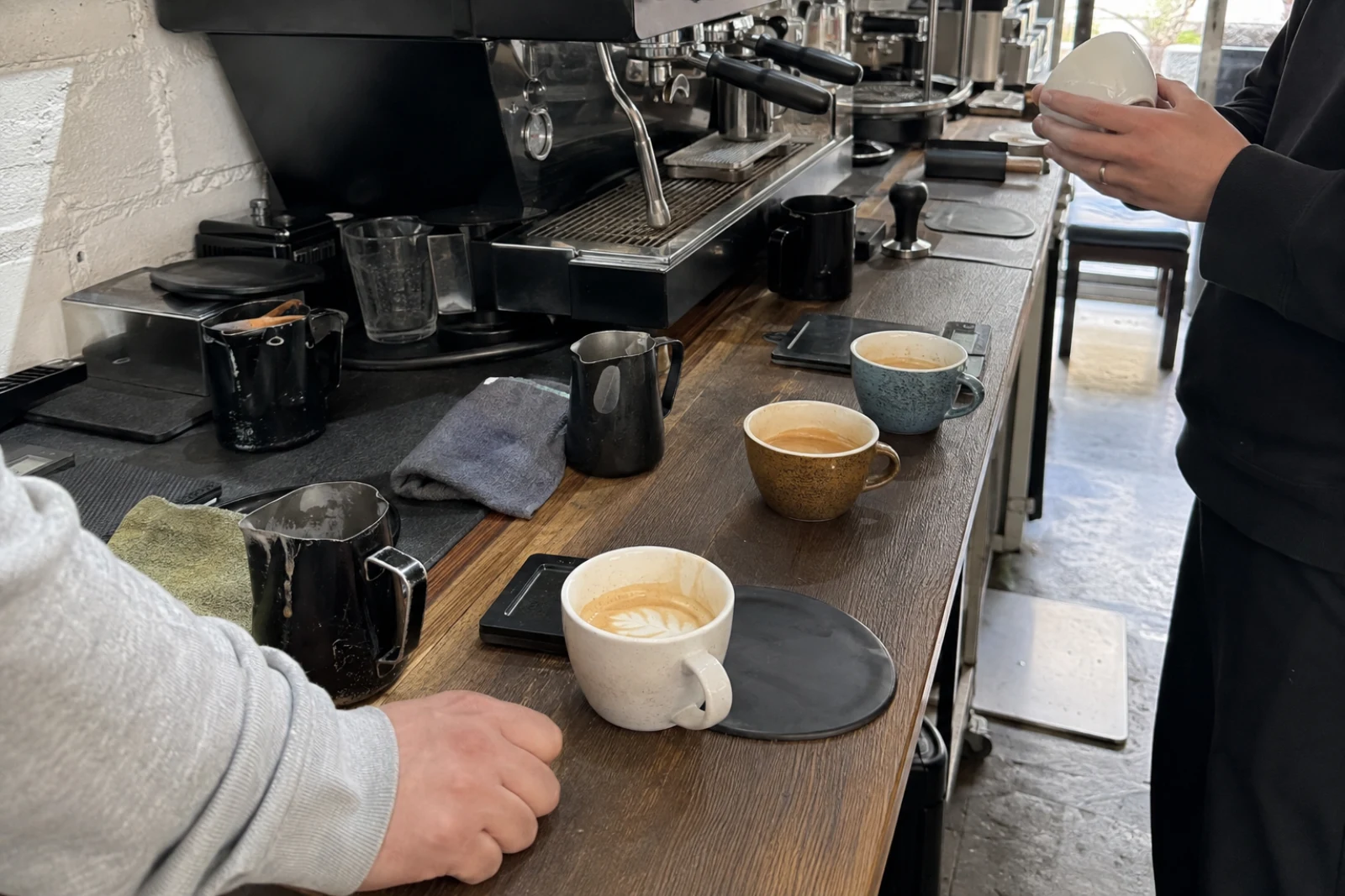 Used milk pitchers and practice cups lined up beside an espresso machine during a latte art workshop