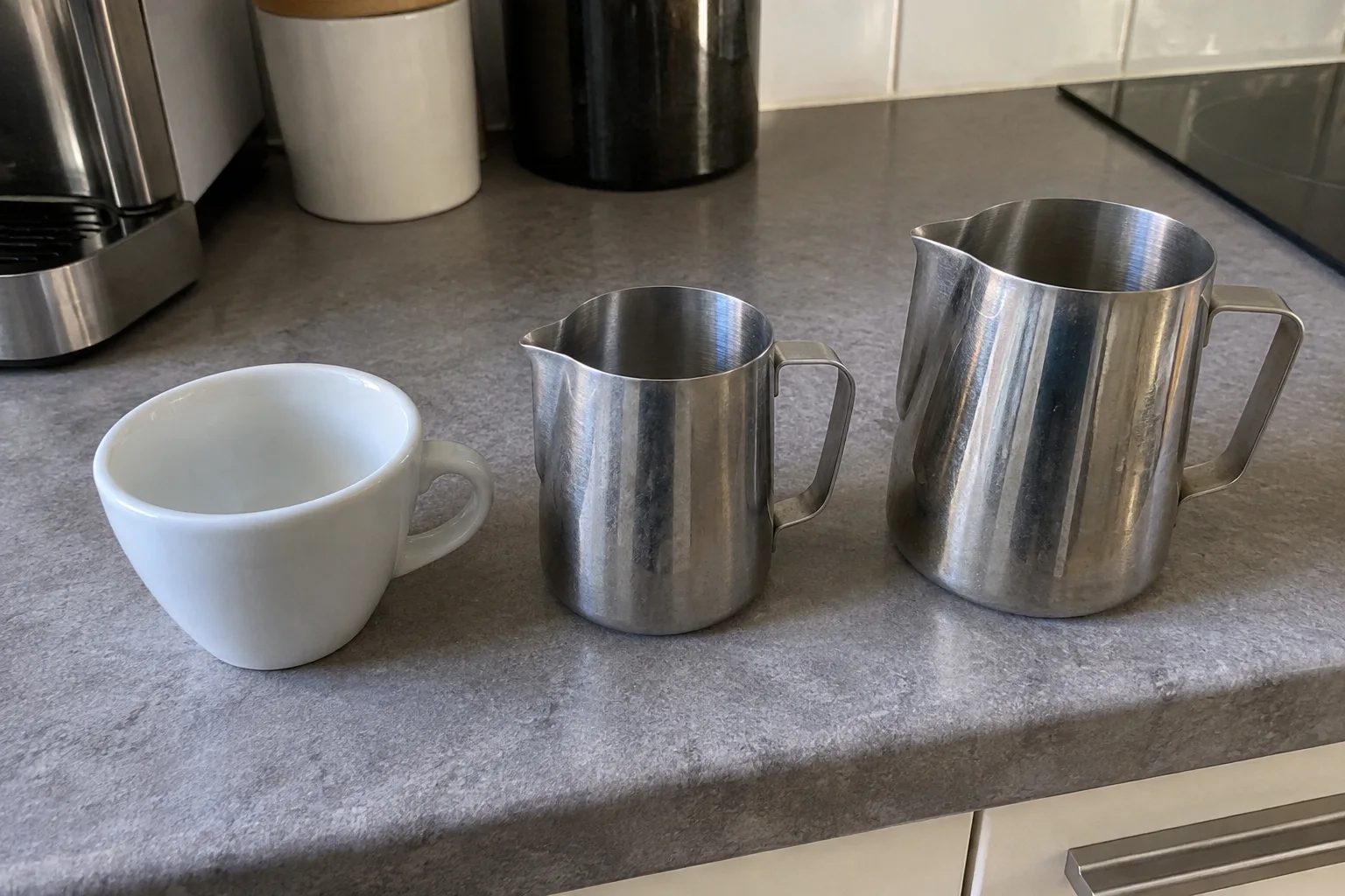 A small and a large stainless steel milk pitcher beside an empty coffee cup